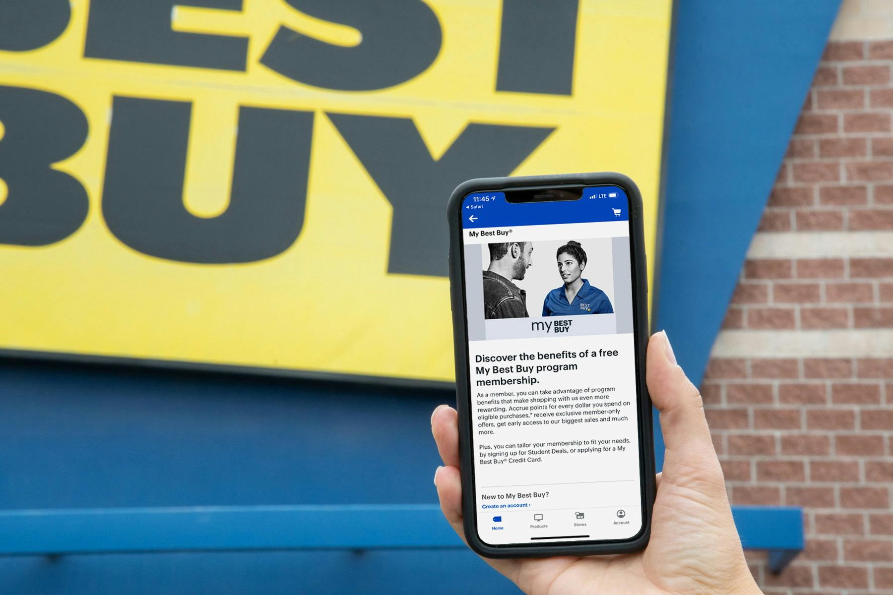 A person's hand holding up an iPhone displaying the My Best Buy program membership page in the Best Buy app in front of a Best Buy store.
