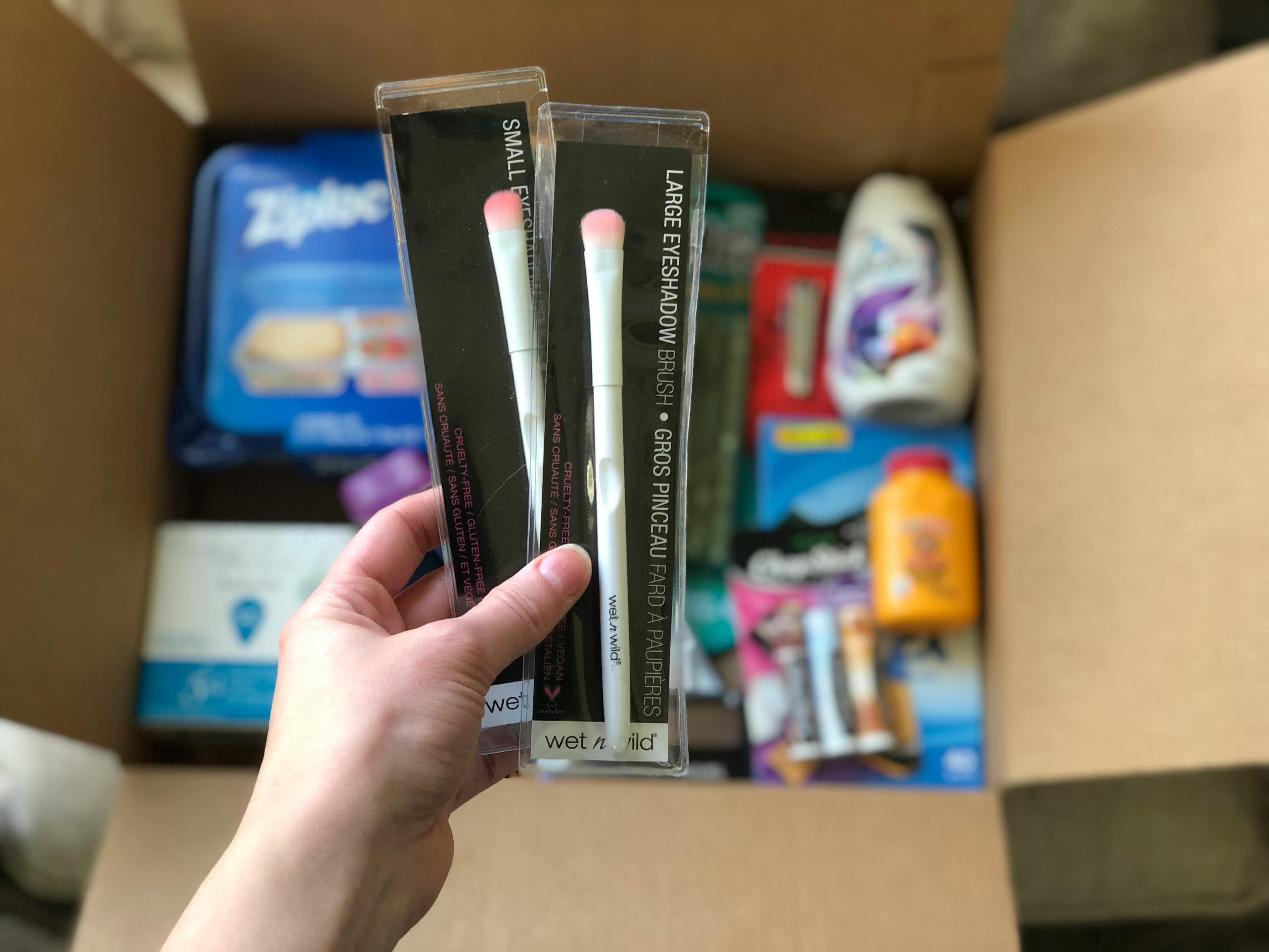 Hand holding Amazon Subscribe & Save items up with a box of household items in the background.