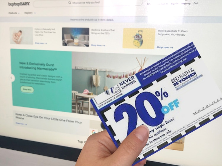 a woman's hand holds a coupon for Bed Bath and Beyond in front of a computer monitor showing the website Buy Buy Baby.