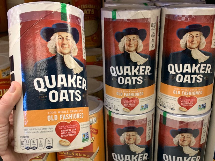 A person holding a canister of Quaker Oats Quick Oatmeal in front of a store shelf with other canisters of Quaker Oats Oatmeal.