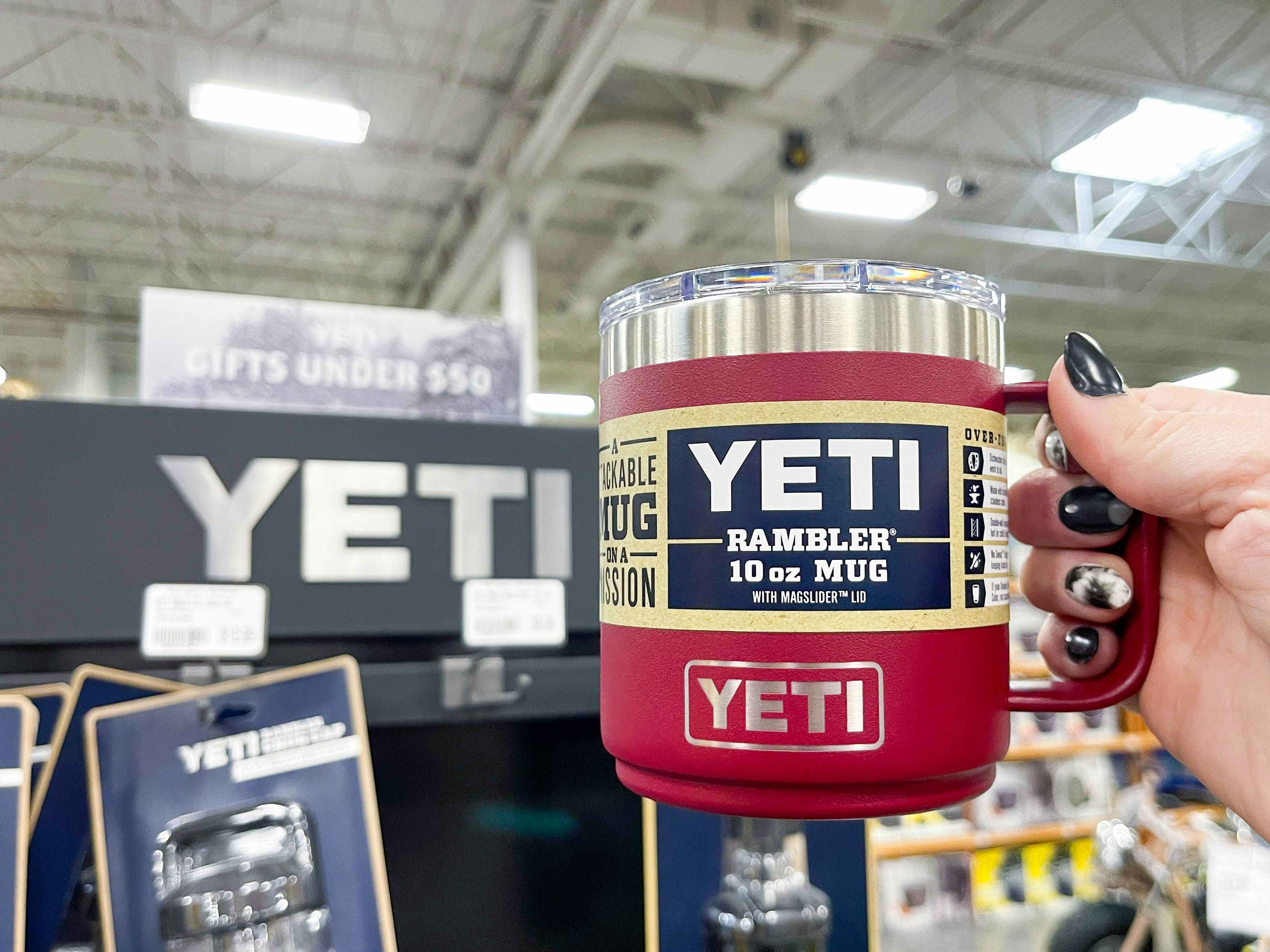 A person's hand holding a Yeti Rambler mug next to a Yeti display inside a store.