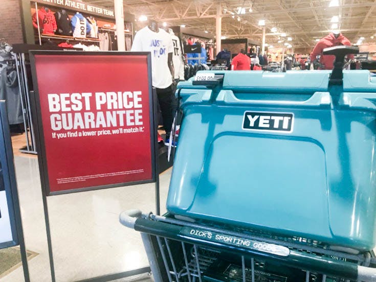 A Yeti cooler sitting in a Dick's Sporting Goods shopping cart next to a sign that reads, "Best Price Guarantee. If you find a lower price, we'll match it." inside a Dick's store.