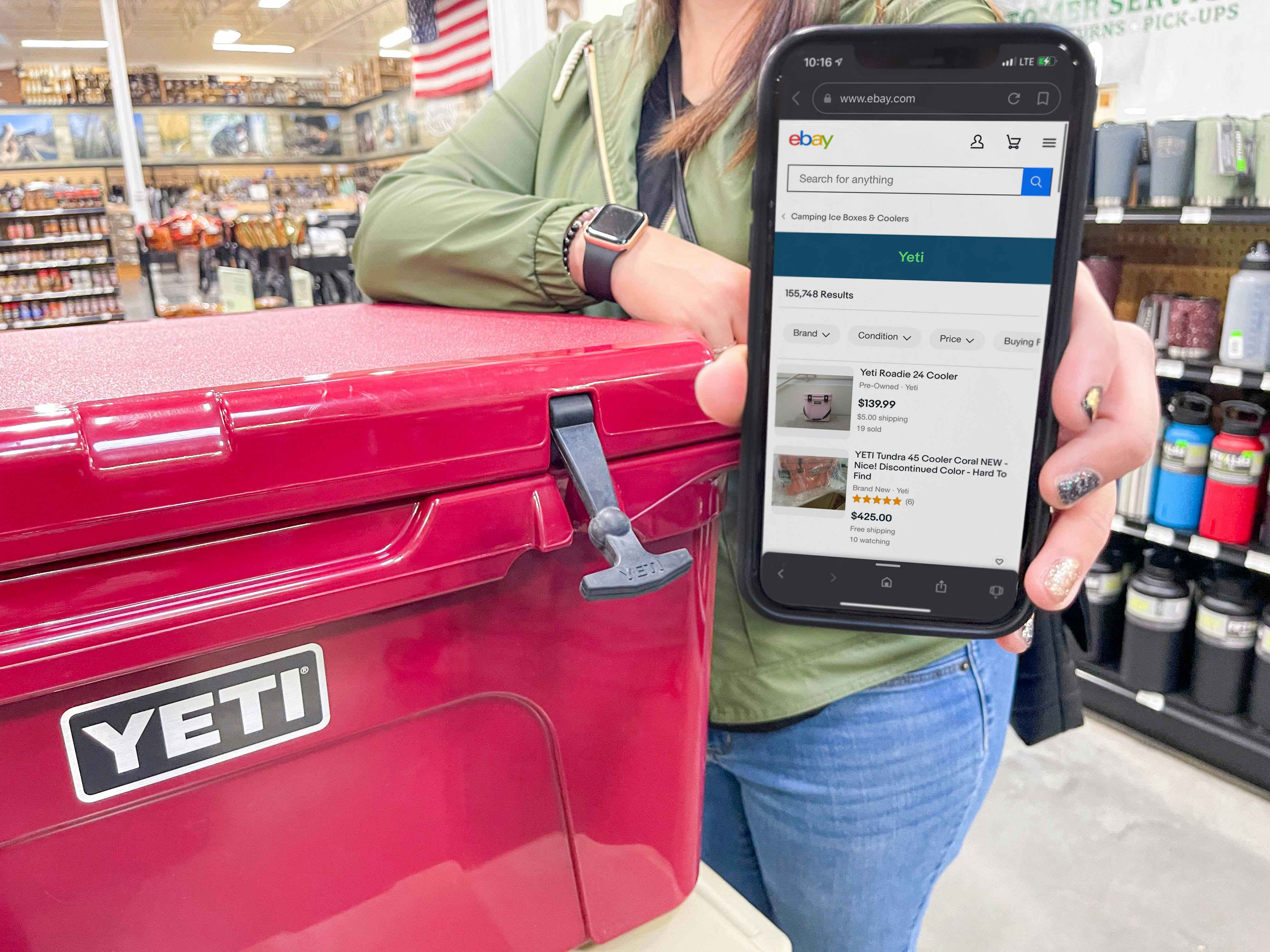 A person resting their elbow on a Yeti cooler and holding up their iPhone displaying the search results for Yeti on the eBay app.