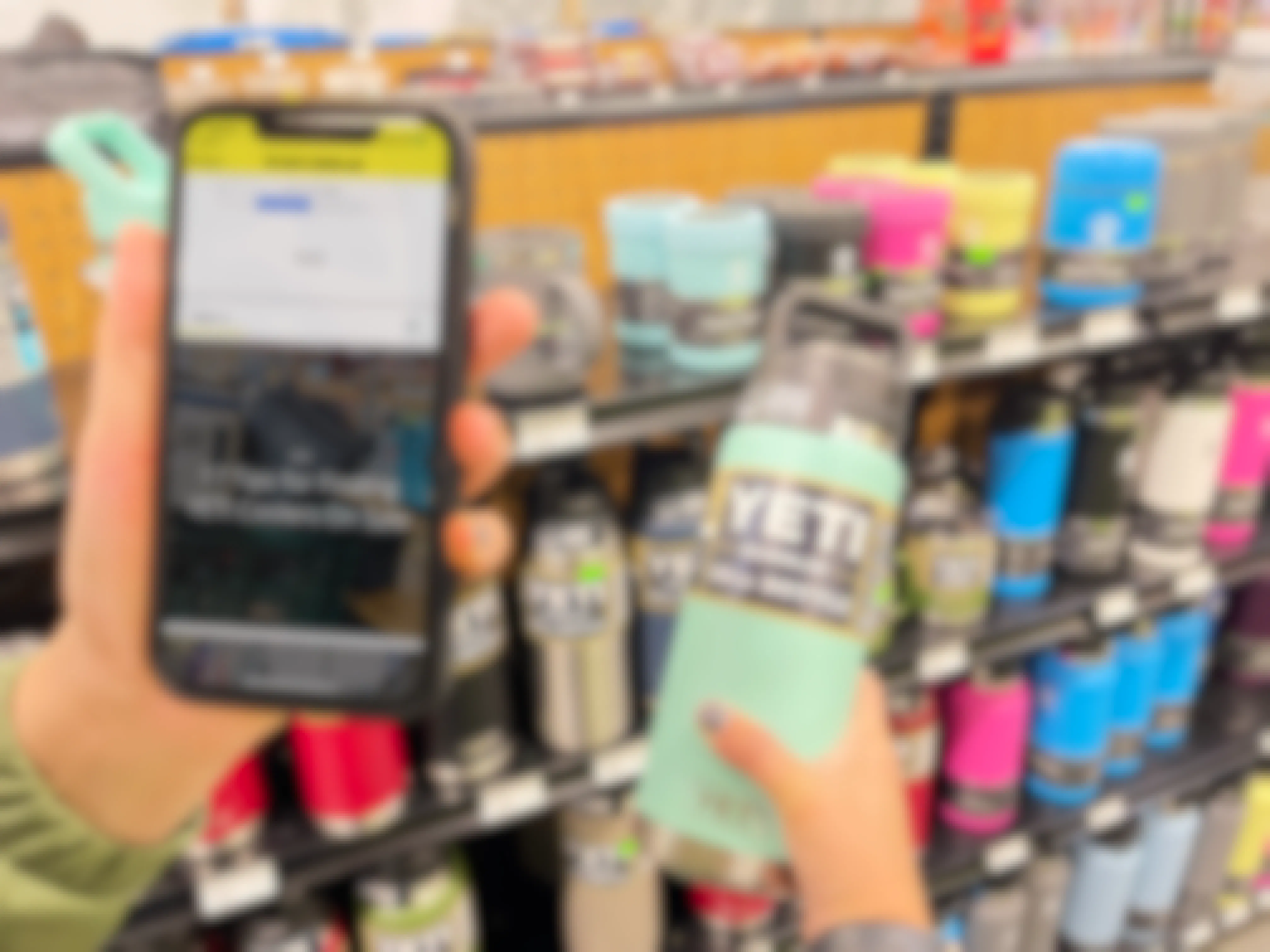 A person's hand holding up their cell phone with the KCL app open to Yeti tips and their other hand holding a Yeti Rambler water bottle in front of a shelf of Yeti products.