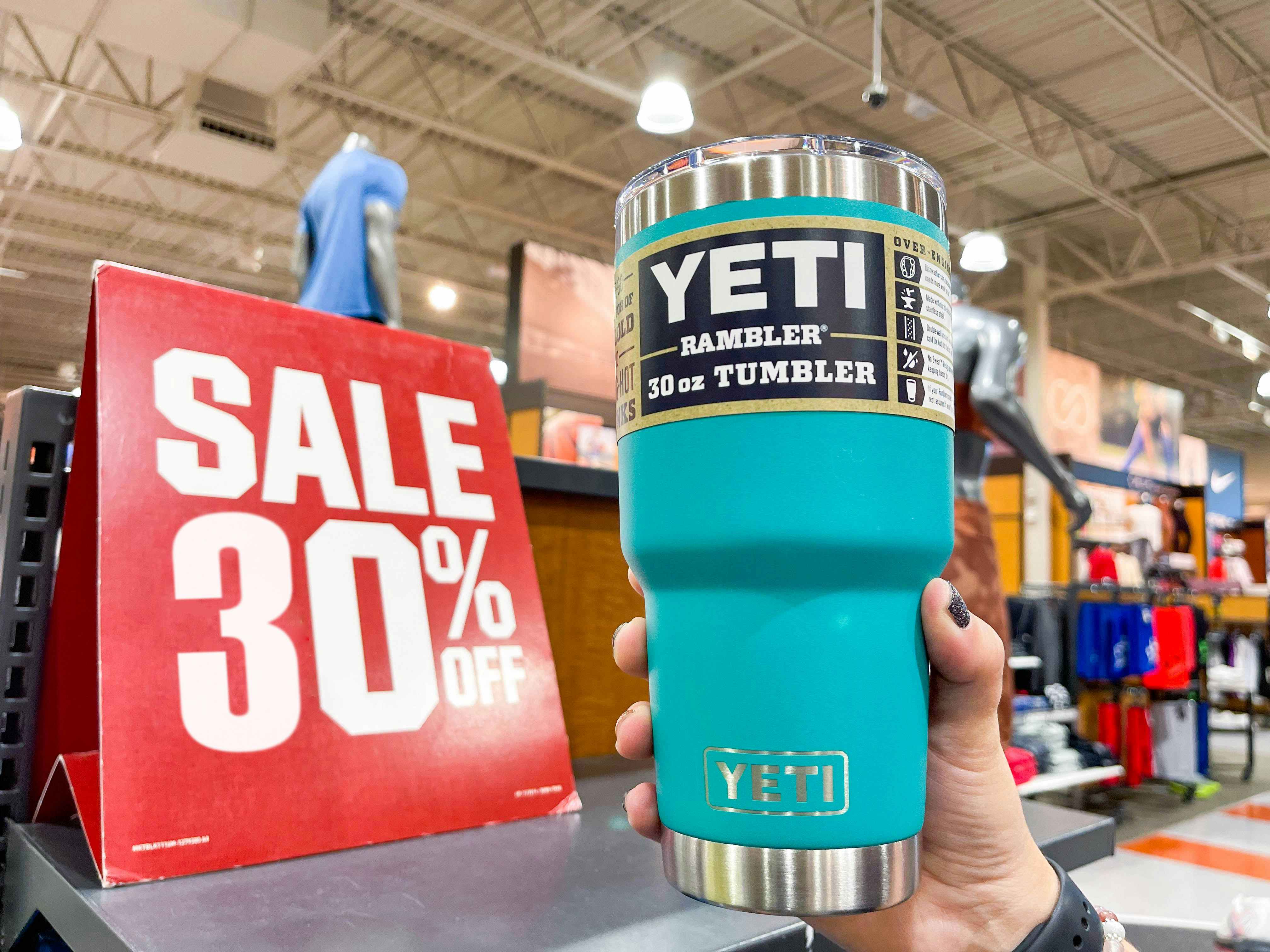 A person's hand holding a Yeti Rambler tumbler near a 30% off sale sign.