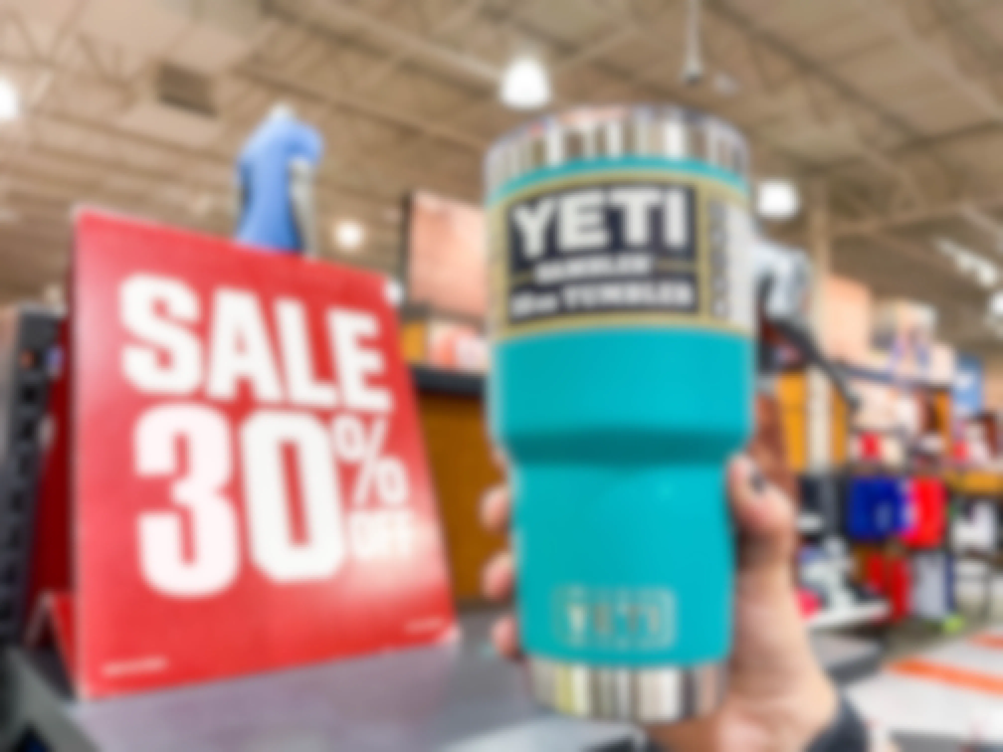 A person's hand holding a Yeti Rambler tumbler near a 30% off sale sign.