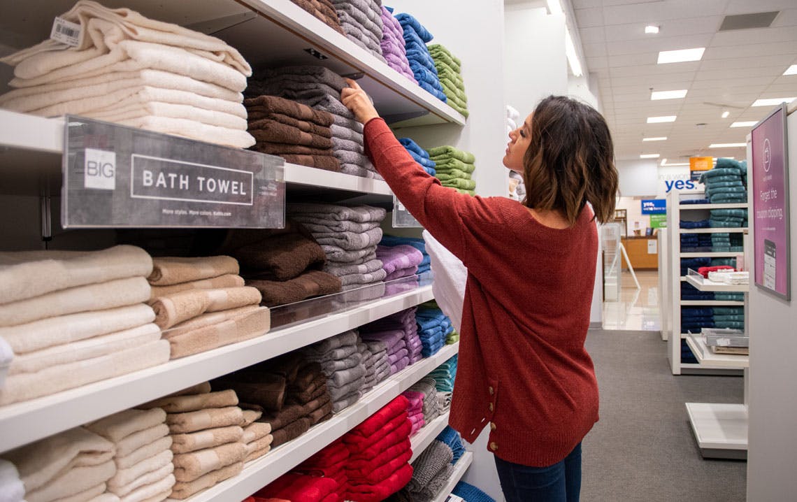 A woman in the bath section at Kohl's, reaching for a The Big One brand towel from a wall of towels.