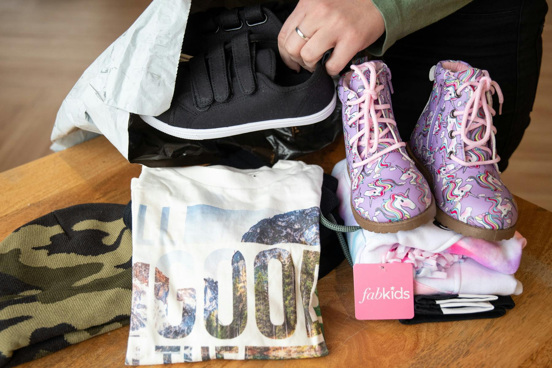 FabKids brand clothing: Purple unicorn print boots, folded t-shirt with words on it, camo beanie and black velcro sneakers