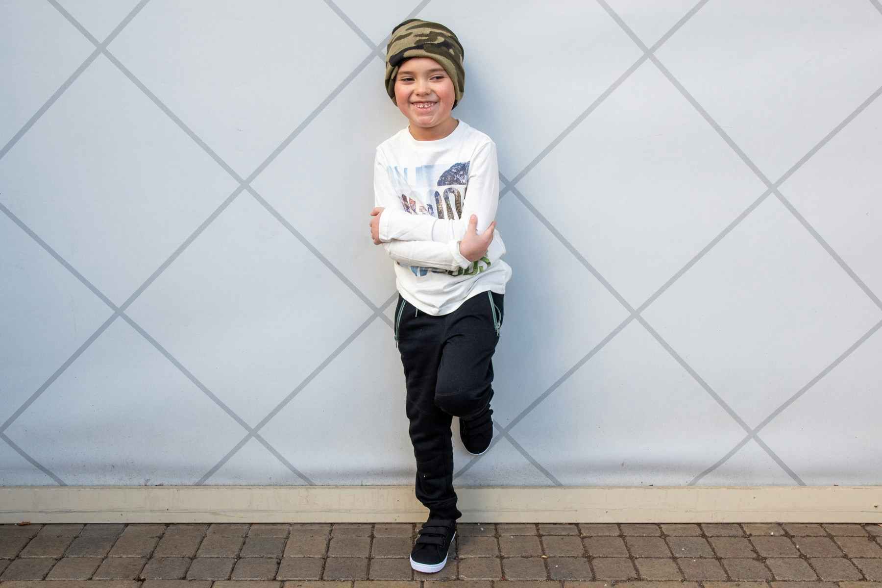 Boy standing against a wall with one leg up on wall behind him smiling wearing FabKids Wild Woods Outfit, Long Sleeve t-shirt, black joggers, black tennis shoes and camo beanie FabKids