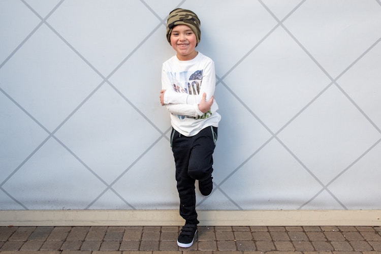 Boy standing against a wall with one leg up on wall behind him smiling wearing FabKids Wild Woods Outfit, Long Sleeve t-shirt, black joggers, black tennis shoes and camo beanie FabKids