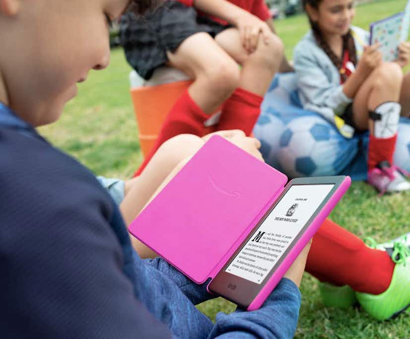 A child in soccer gear holding an Amazon Kindle displaying the beginning of a book chapter with other children with soccer gear looking at another Kindle in the background.