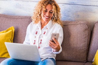 A woman sitting on a couch using a laptop and looking at her phone.