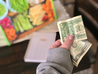 A hand holds cash while a planner and grocery bag are in the background.