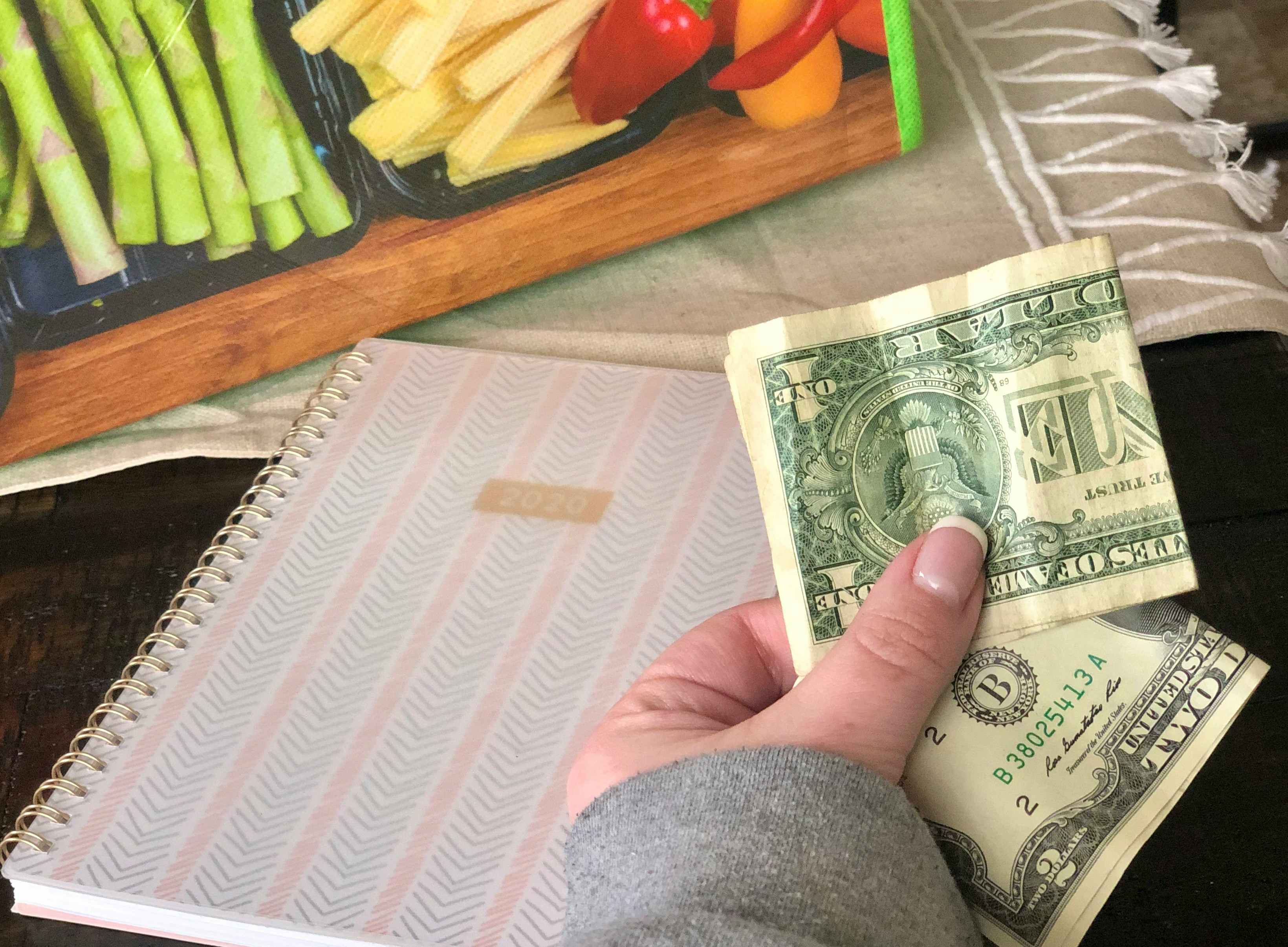 A hand holds cash while a planner and grocery bag are in the background.
