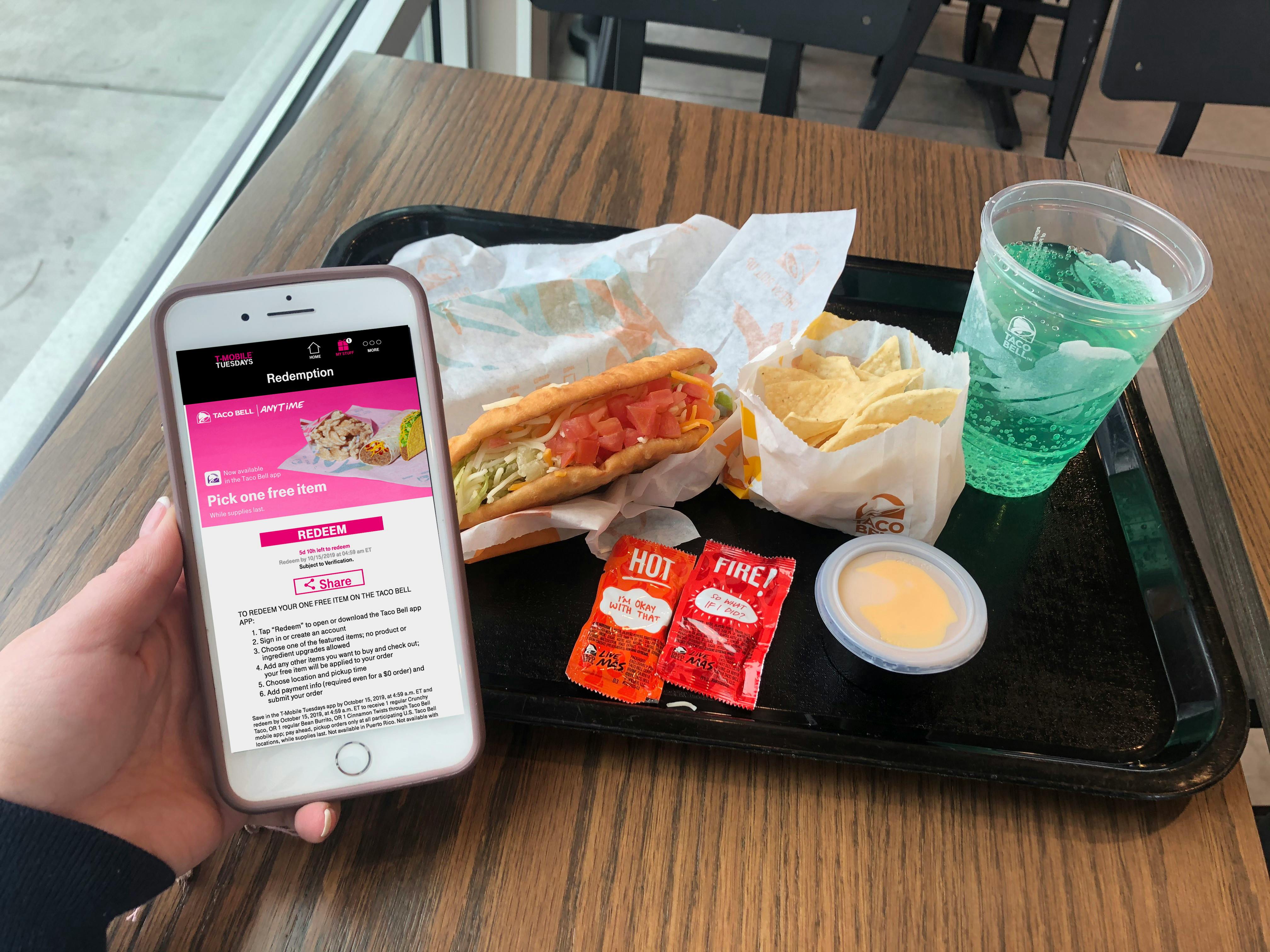 A person's hand holding a cell phone displaying the T-Mobile Tuesdays app's offer redemption page for a free Taco Bell item with Taco Bell food on a tray in the background.