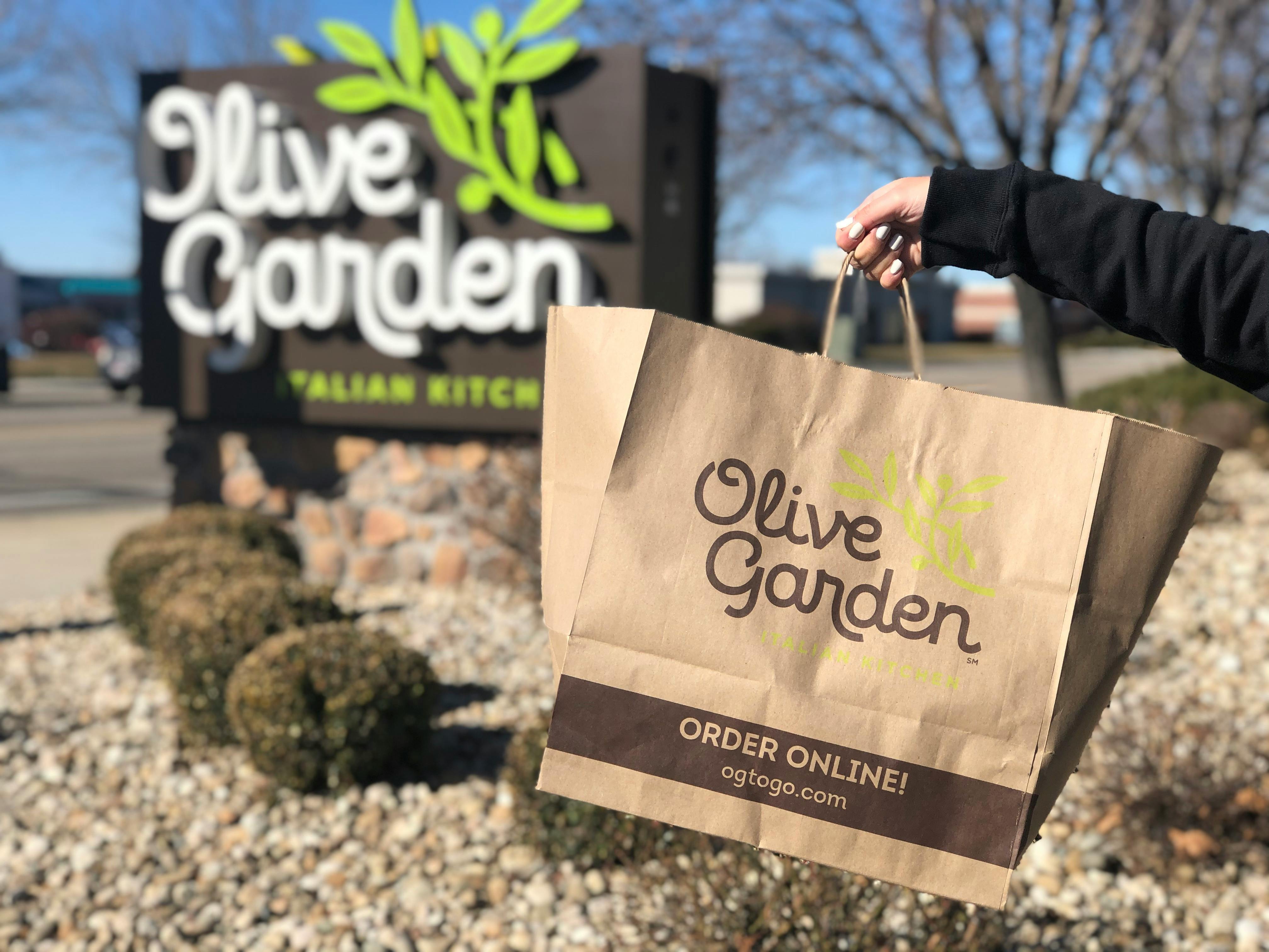 a bag of to go olive garden being held out in front of outside olive garden sign 