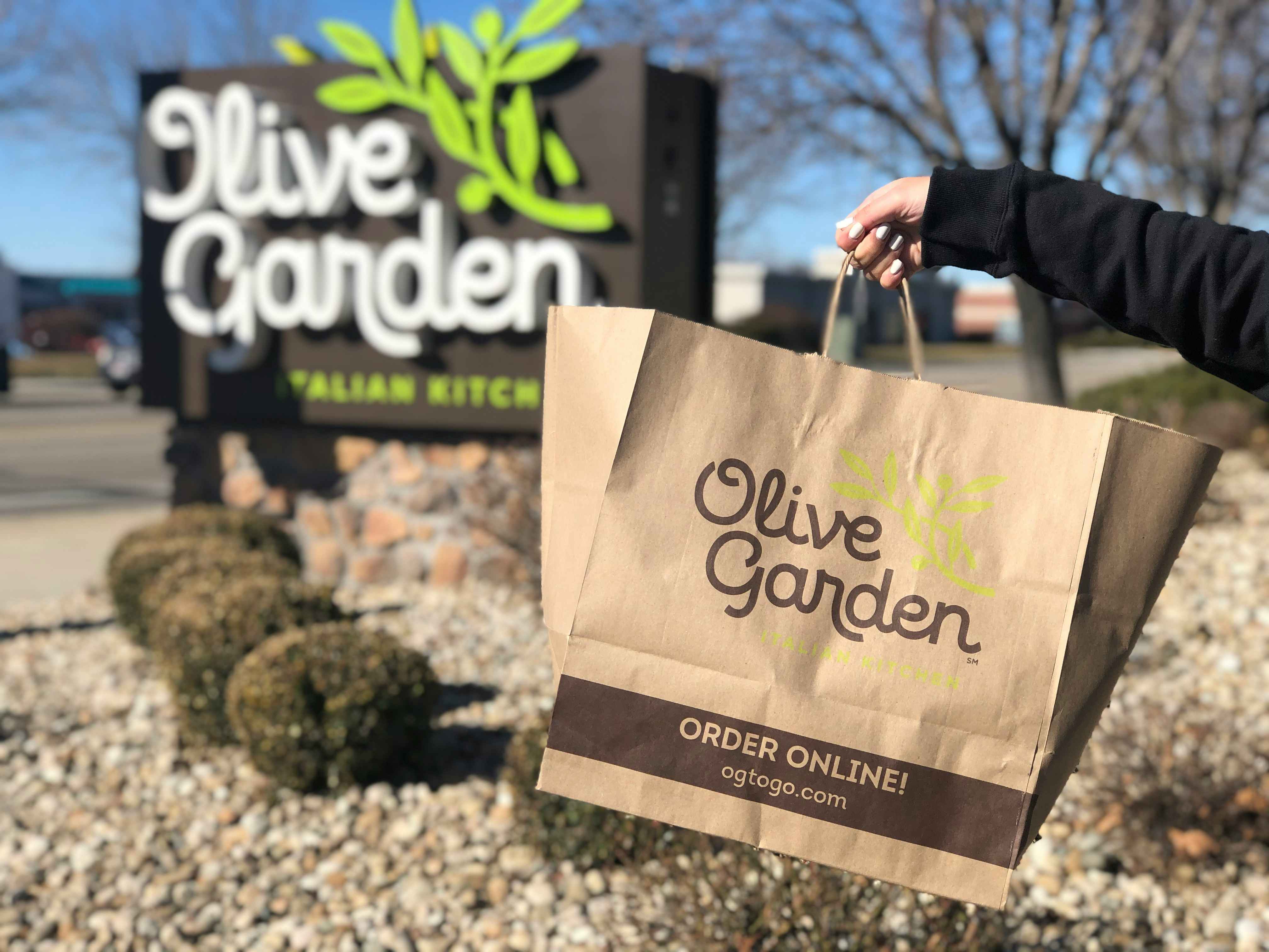 Someone holding an Olive Garden to go bag in front of an Olive Garden sign