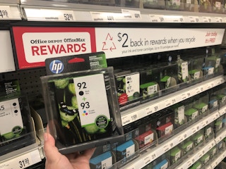 A person holding a package of printer ink cartridges in front of a shelf with a sign that says, "$2 back in rewards when you recycle.