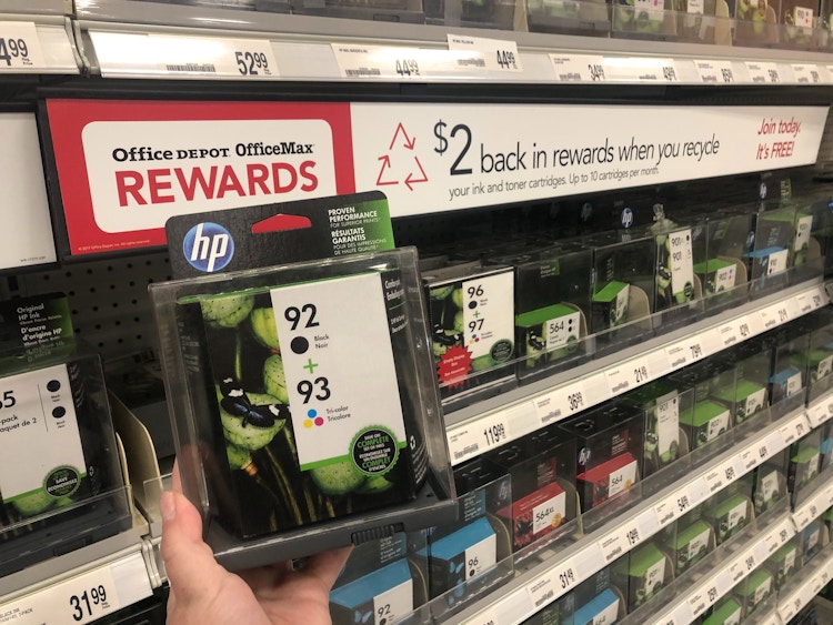 A person holding a package of printer ink cartridges in front of a shelf with a sign that says, "$2 back in rewards when you recycle.