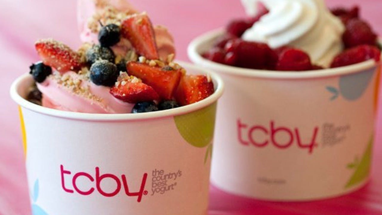 Two cups of frozen yogurt from TCBY with fruit toppings sitting next to each other on a pink table.
