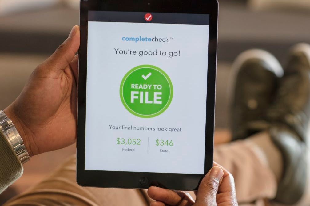 A person sitting down, holding up an iPad displaying the TurboTax completecheck screen showing that the user's tax forms are ready to be filed.