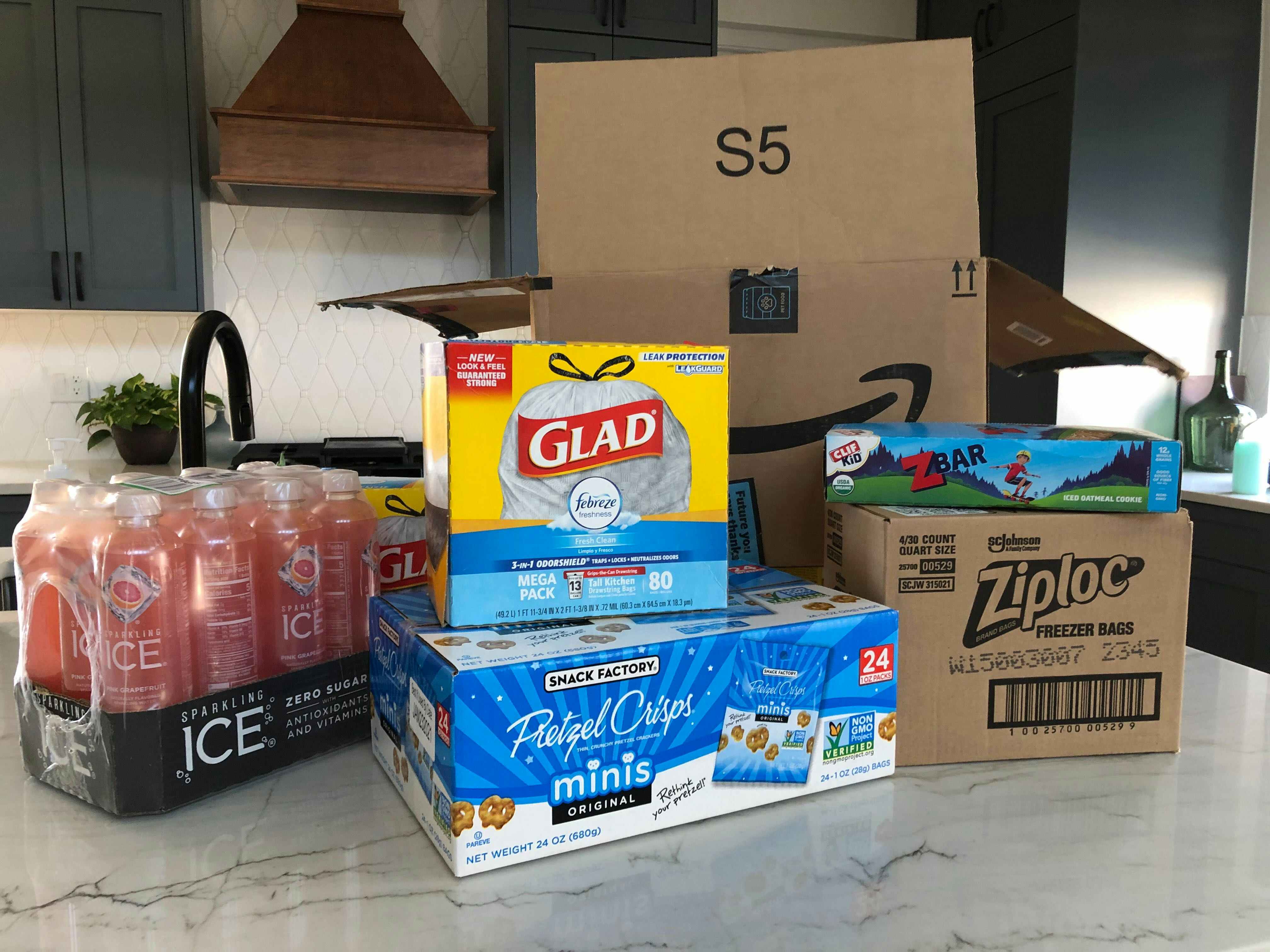 Glad garbage bags, sparking juice, pretzel snack bags, ziplock bags, and zbars sitting on a counter in front of an Amazon box
