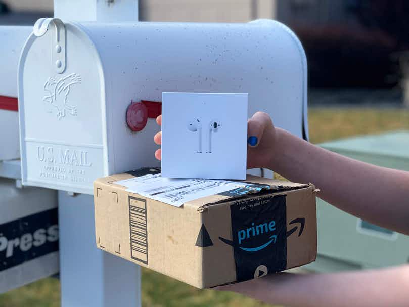 A person holding a box of Apple Airpods and an Amazon Prime delivery box near a mailbox.