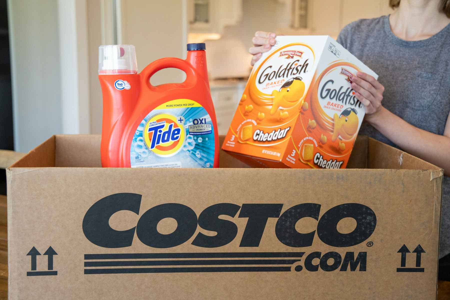 A woman pulling gold fish crackers and tide from a Costco.com shipping box.