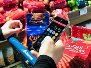 Person shopping the produce aisle, close up of her hands pushing cart, using iPhone calculator