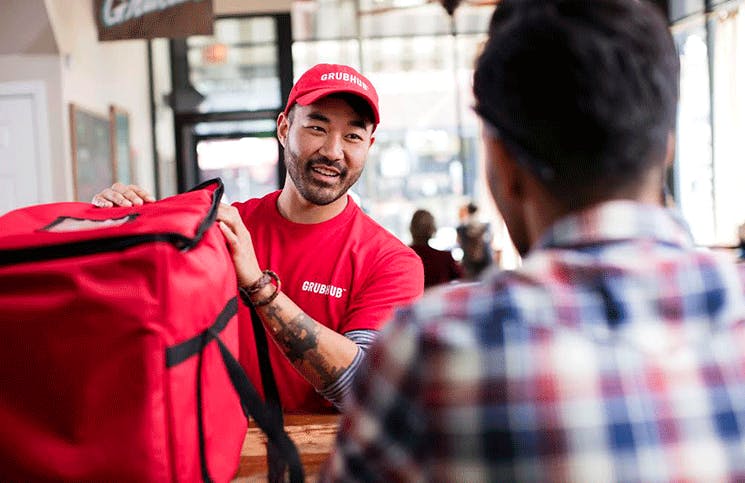 - A person wearing a Grubhub hat and t-shirt setting a food delivery bag on the counter in front of a customer. The customer is out of focus in the foreground, facing the Grubhub worker.