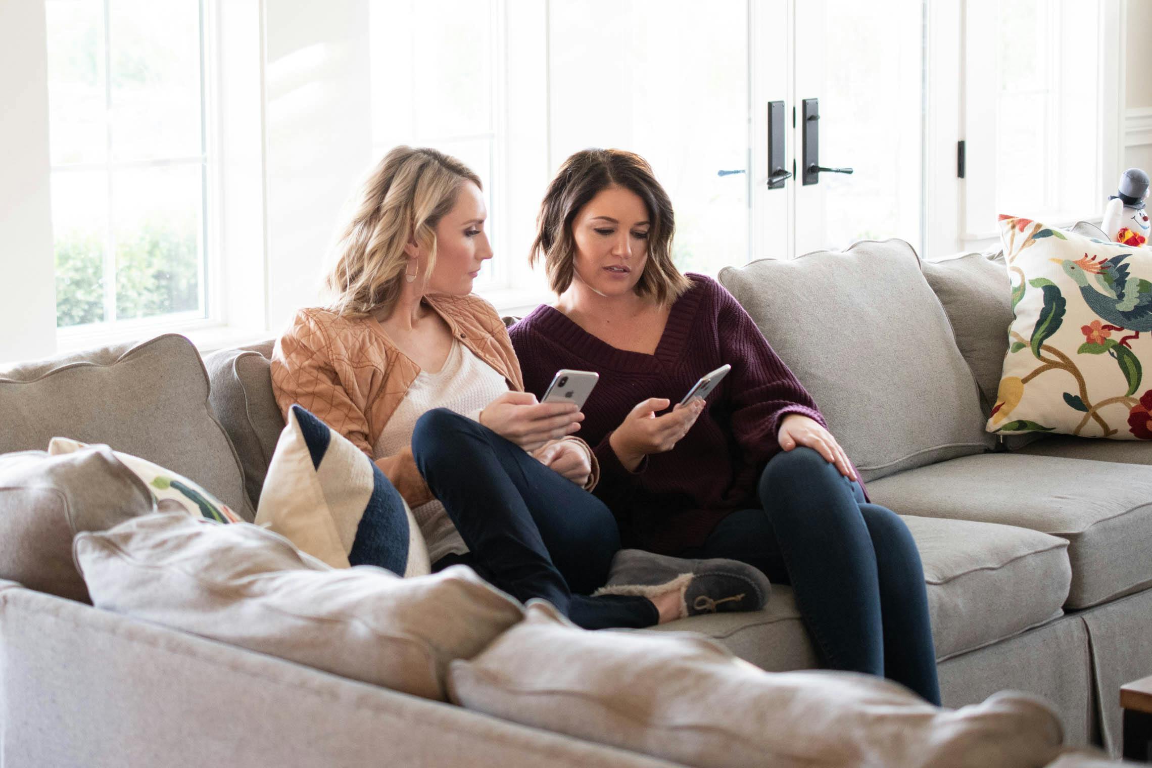 Two women sitting next to each other on a sofa while looking at their cell phones.