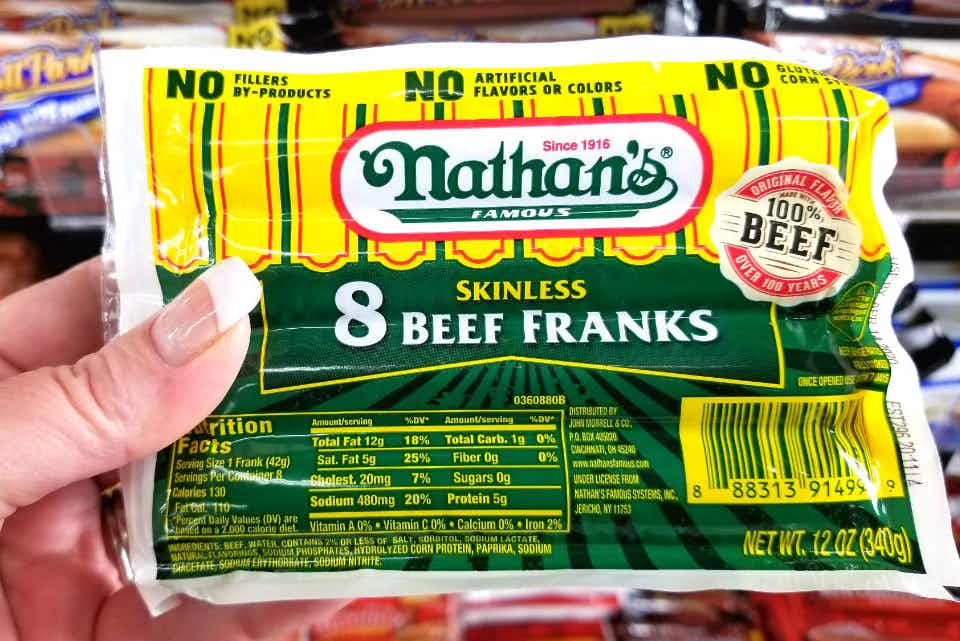 A person's hand holding up a package of Nathan's Famous skinless beef franks in front of the hot dog shelf at a grocery store.