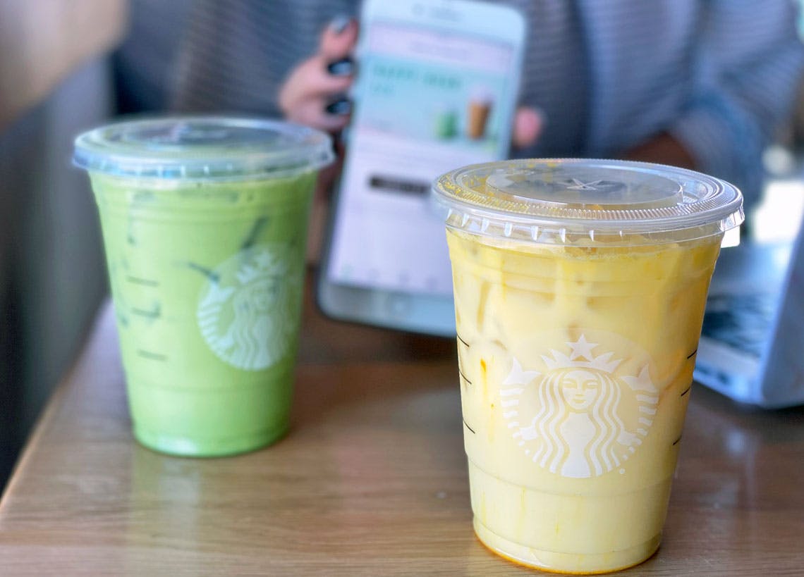 Two matcha Starbucks drinks sitting on a table in front of a person holding their phone.