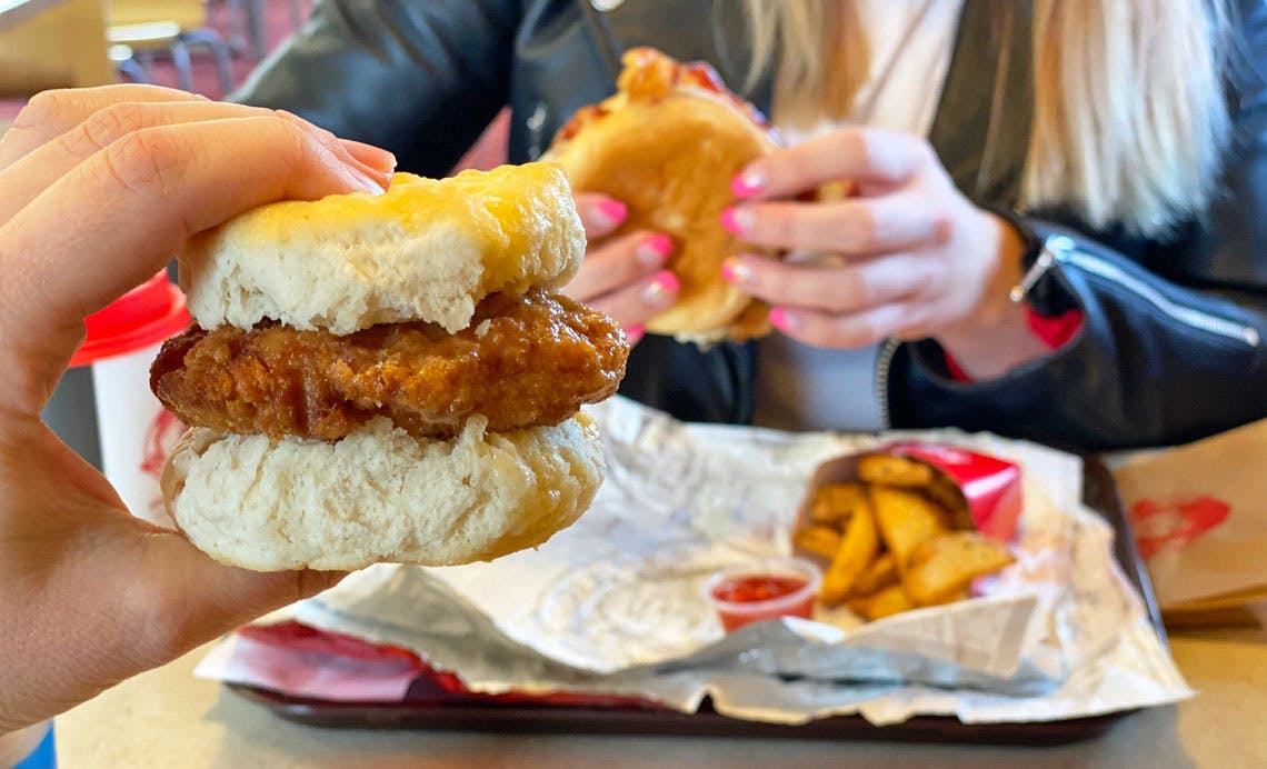 A person's hand holding a Wendy's breakfast sandwich up in front of another person eating a Wendy's meal in a booth at Wendy's.