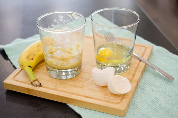 cutting-board-with-a-glass-with-mashed-bananas-and-another-glass-with-a-raw-egg-in-it