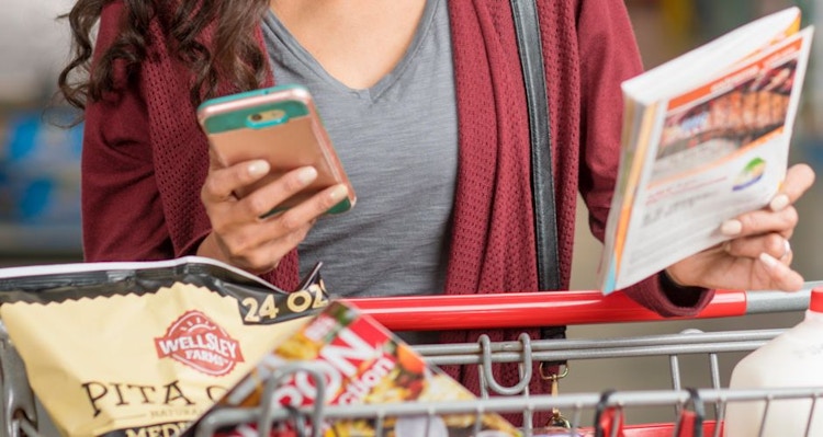 woman holding a cellphone and BJ's Wholesale coupon book while pushing a shopping cart