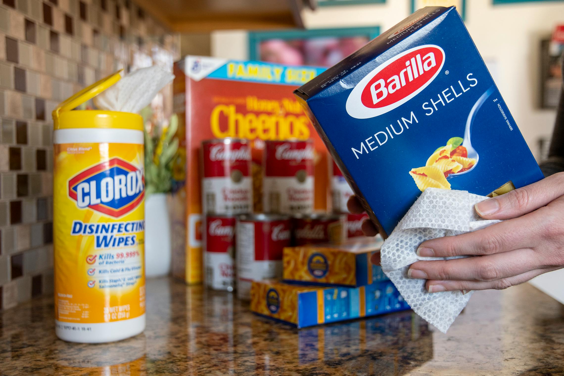 A person cleaning a box of Barilla medium shell past with a Clorox wipe with other groceries piled on the counter behind.