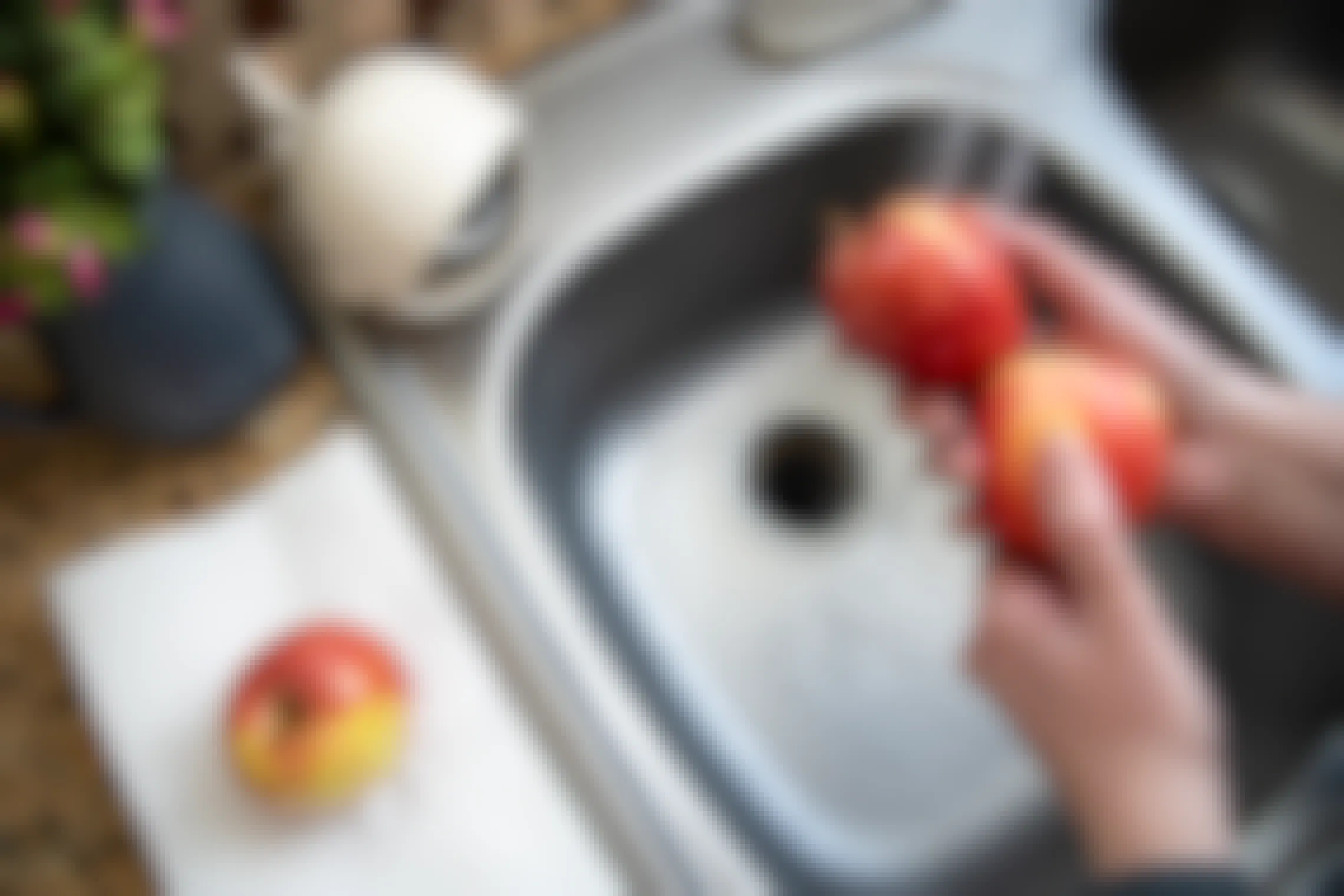 Two apples being washed over a kitchen sink with a third sitting on paper towel next to it.