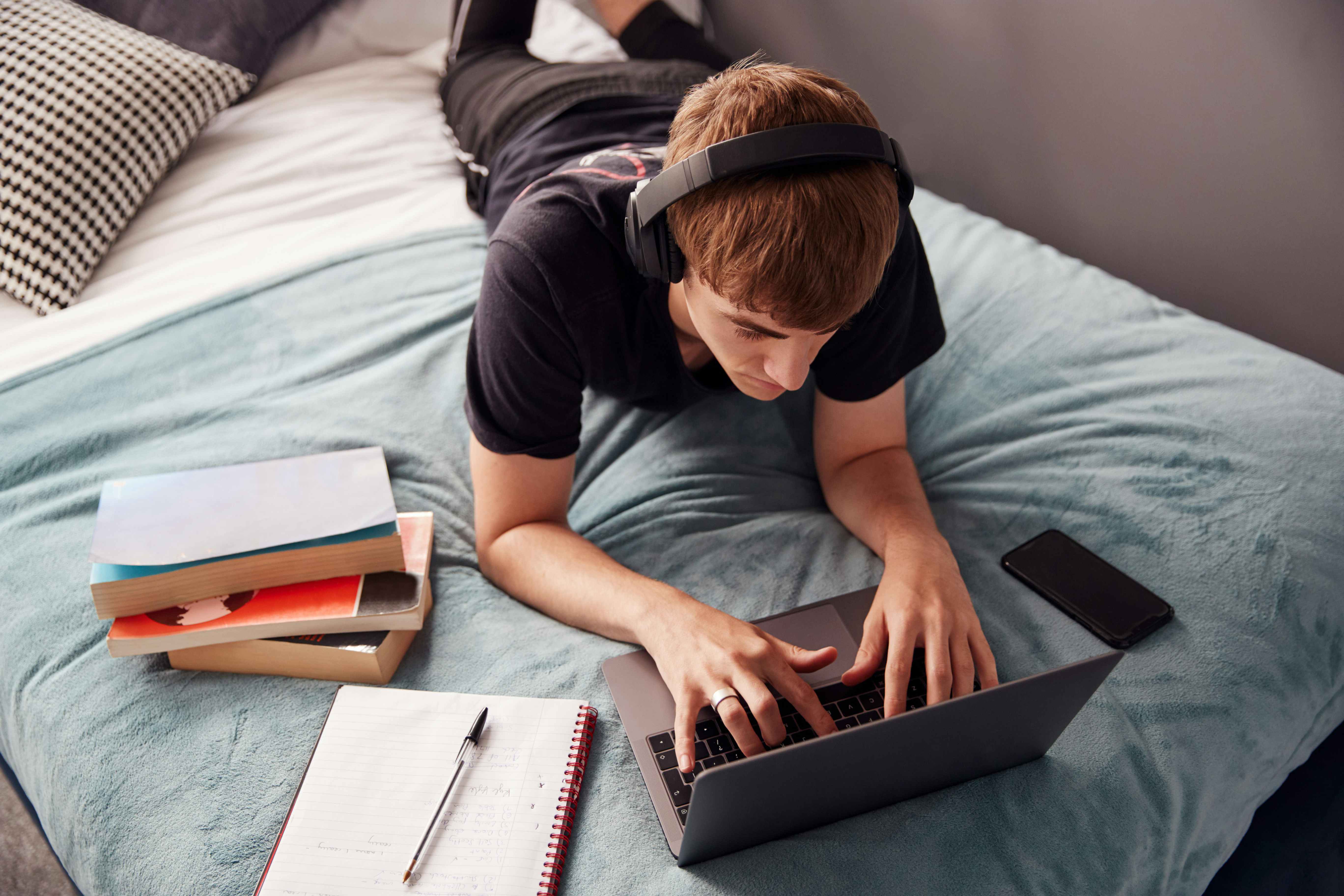 A student with headphones lying down, using a laptop on a bed next to some books, a notebook, and a phone.