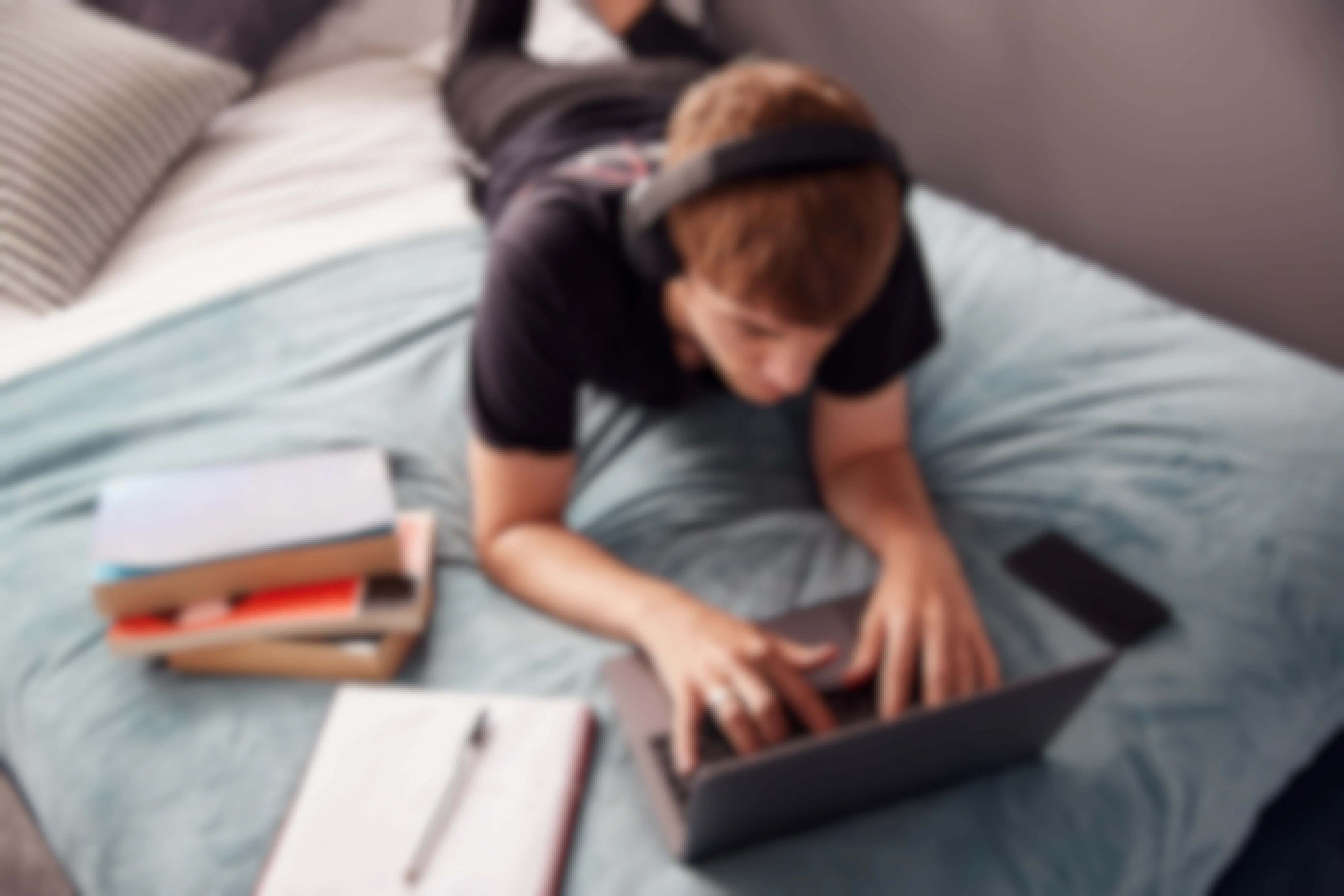 A student with headphones lying down, using a laptop on a bed next to some books, a notebook, and a phone.