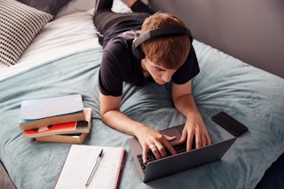 A student with headphones lying down, using a laptop on a bed next to some books, a notebook, and a phone.