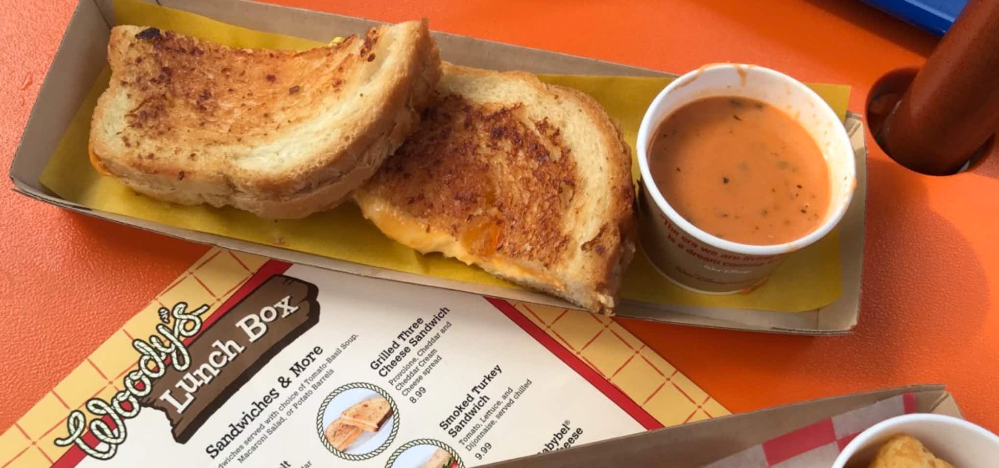 Grilled cheese on a plate with tomato soup with a Woody's Lunch Box menu nearby
