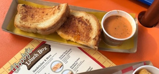 Grilled cheese on a plate with tomato soup with a Woody's Lunch Box menu nearby