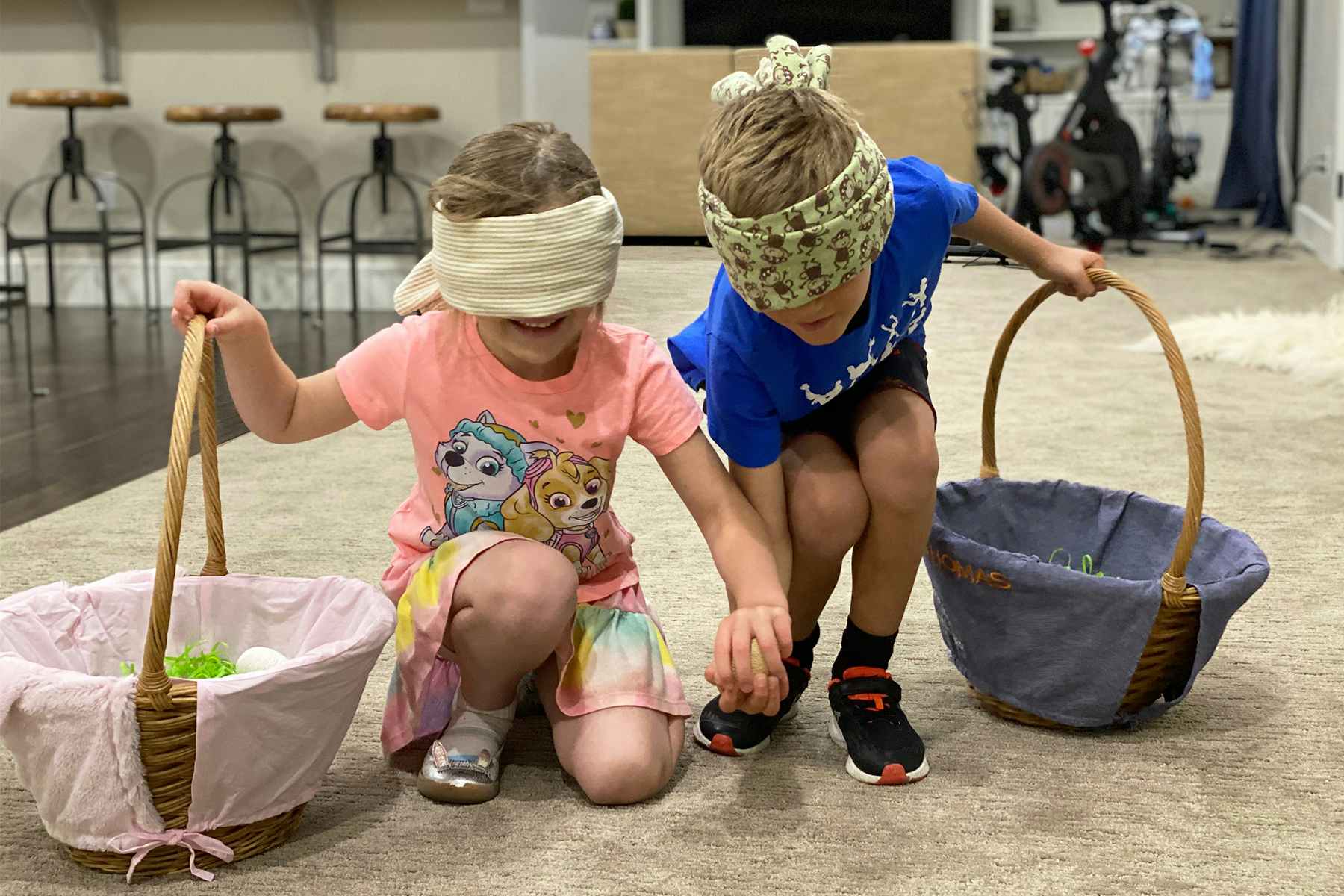 Two kids holding wearing blindfolds, holding Easter baskets, collecting Easter eggs inside a house.