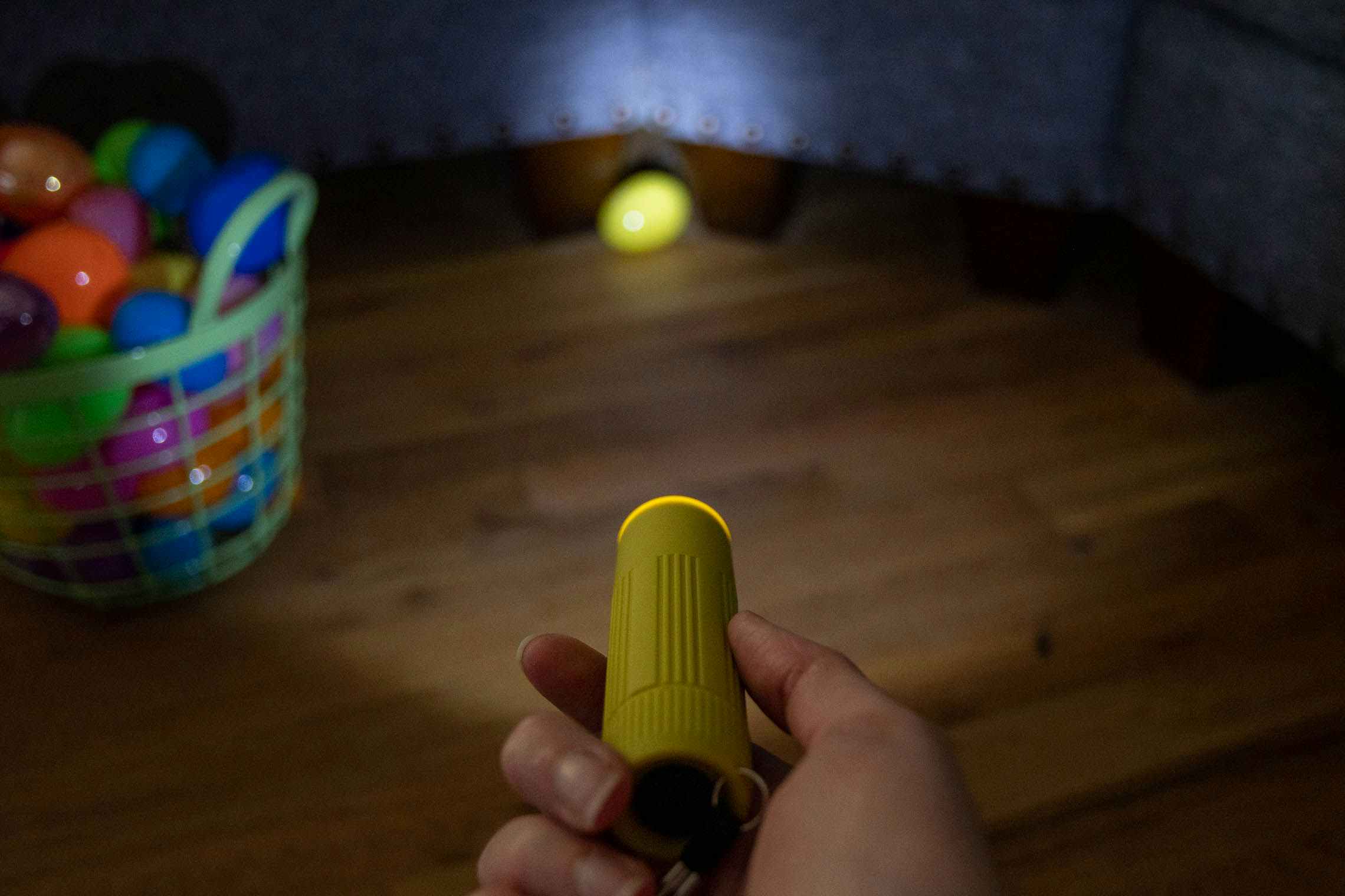 A flashlight pointed on a plastic easter egg with a basket of plastic eggs next to it.