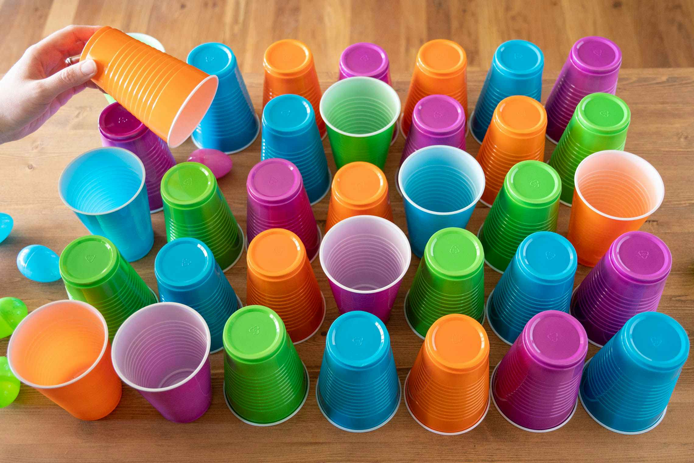 Pink, green, orange, and blue plastic cups set out on a table in a pattern with plastic Easter eggs under them and next to them.