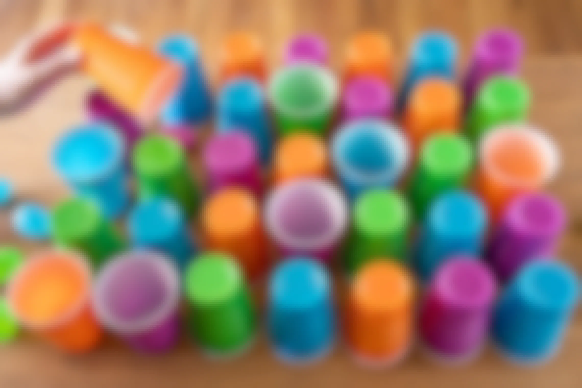 Pink, green, orange, and blue plastic cups set out on a table in a pattern with plastic Easter eggs under them and next to them.