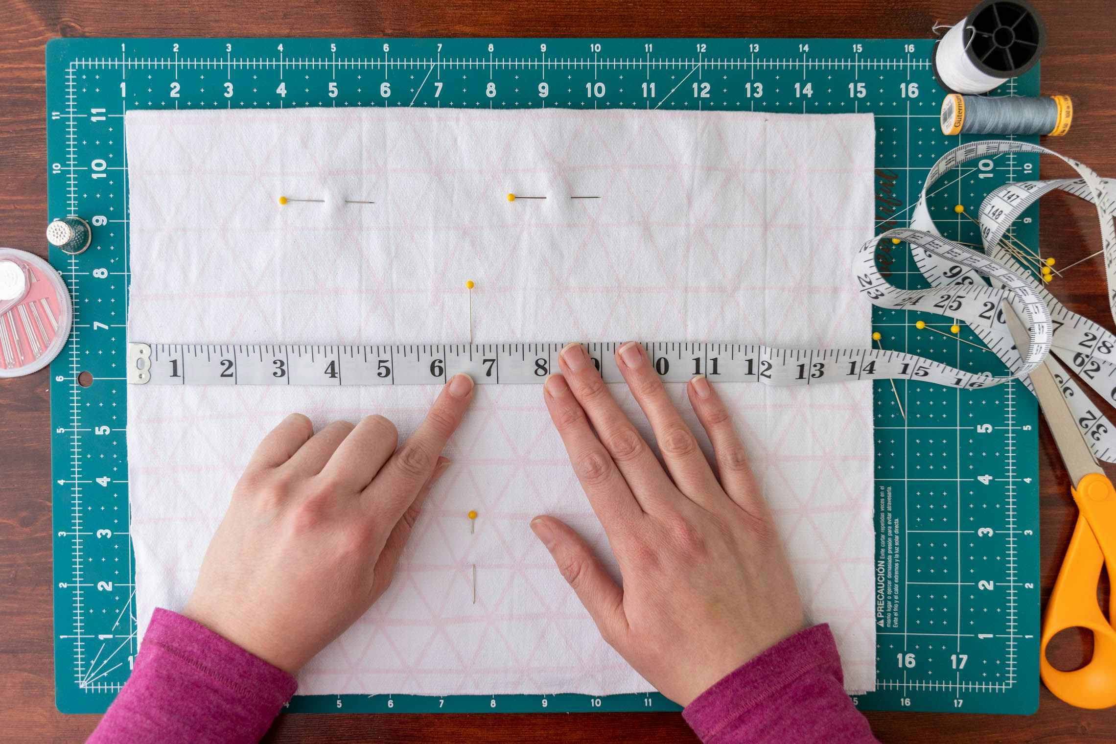 A cutting mat, sewing needles, thread, scissors, and a measuring tape laying on top a piece of fabric. Two hands are positioned on top, pointing to a measurement on the tape measure.