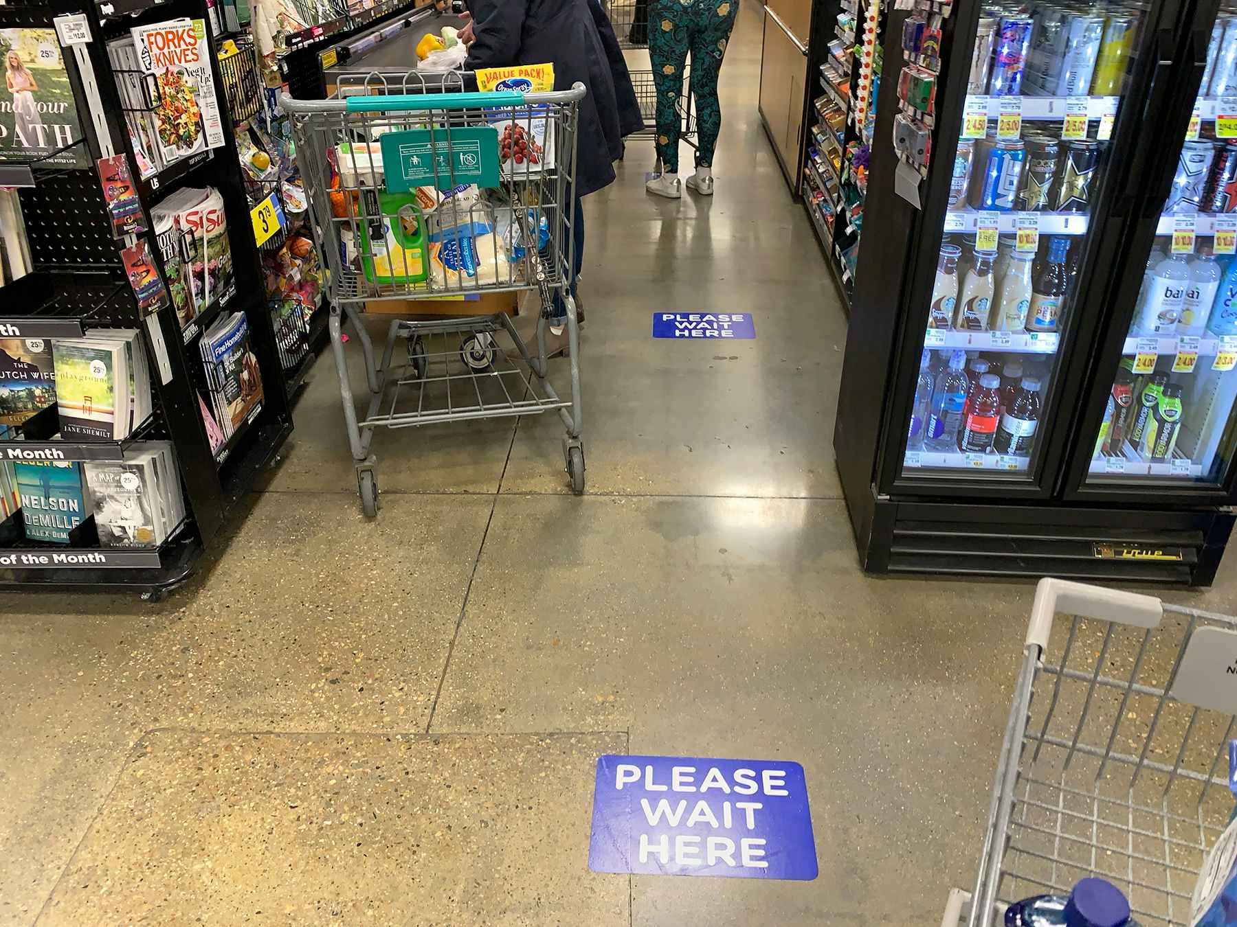 A checkout line with people shopping, standing near markers on the floor that say, "Please Wait here