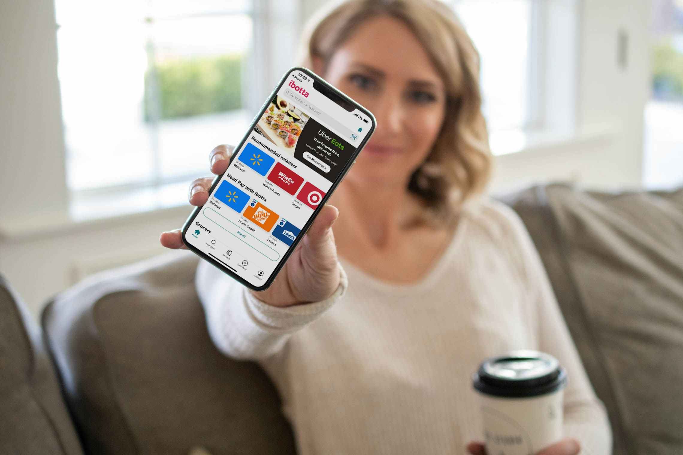 A woman holding a phone out in front of her, facing the camera, with the ibotta app open on it.